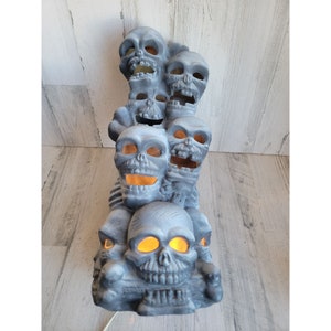 May include: A stack of gray ceramic skulls with glowing orange eyes. The skulls are arranged in a tower formation, with the largest skull at the bottom and the smallest skull at the top. The skulls are all facing forward, and their mouths are open in a menacing grin.