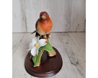 May include: A porcelain figurine of a red-breasted bird perched on a branch with a white flower. The bird is sitting on a wooden base.
