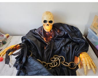 May include: A Halloween decoration of a skeleton wearing a black robe with a chain around it. The skeleton's head is lit up with warm white lights.