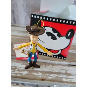 Puede incluir: Un adorno de plástico de Woody de Toy Story, con su característico sombrero de vaquero y chaleco. Lleva un cuerno dorado y tiene una expresión amistosa en su rostro. El adorno está empaquetado en una caja roja con una imagen de Mickey Mouse.
