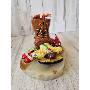 May include: A hand-painted ceramic figurine of a clown stuck upside down in a brown boot. The clown is wearing a yellow and red costume and a red hat. The figurine is sitting on a white stone base.
