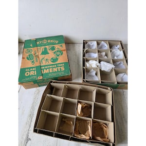 Könnte beinhalten: Ein vintage Shiny Brite Pappkarton für gläserne Weihnachtsbaumschmuck-Kugeln mit grünen und beigen Grafiken, inklusive des Textes Shiny Brite Glass Christmas Tree Ornaments. Zwei offene Papp-Einsätze sind vorhanden, einer enthält weißes Papier.