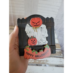May include: A wooden Halloween decoration with a ghost holding a pumpkin and skulls. The decoration has a black picket fence and the words "Happy Halloween" on the bottom.