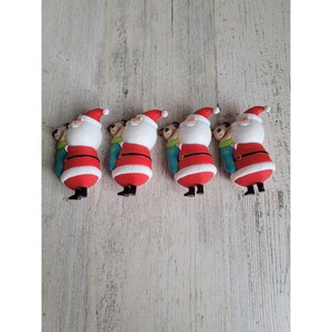 Puede incluir: Cuatro pequeñas figuras de Papá Noel rojas y blancas que sostienen un saco azul y verde. Cada Papá Noel lleva un gorro rojo con ribete blanco y una barba blanca.