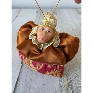 May include: A whimsical monkey ornament with a detailed face, wearing a gold-trimmed hat and collar. The monkey is dressed in a brown satin outfit with a burgundy and gold patterned base. A gold string is attached for hanging.