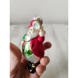 May include: A glass Christmas bauble featuring Santa Claus wearing a red coat and green waistcoat, holding a glass of beer in his right hand.