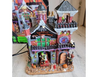 Op de afbeelding: Een gedetailleerd miniatuurgebouw met Halloween-thema, de Spookytown Fire Department, met een zwart dak en een toren. Het gebouw is geschilderd in tinten rood, grijs en paars, met groene ramen en decoratieve elementen. De tekst "Spookytown Fire Department" is zichtbaar.