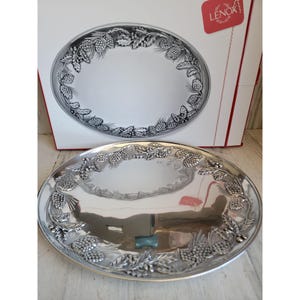 Op de afbeelding: Een zilverkleurige ovale serveerschaal met een gedetailleerde rand van dennenappels en bladeren. De schaal wordt gepresenteerd voor de originele doos, die ook het ontwerp van de schaal heeft. Het Lenox-merkteken is zichtbaar op de doos.