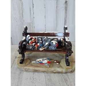 May include: A small, detailed figurine of a clown sleeping on a park bench. The clown is wearing a black and grey coat with white snow on it. The bench is brown with white snow on the top. The figurine is sitting on a white marble base with a brown umbrella lying on the ground.