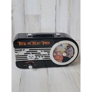 Peut inclure: Radio d'Halloween noir et argent avec un bouton de volume et une image tournante d'une sorcière et de citrouilles. La radio porte l'inscription "Trick or Treat Tunes" sur le devant.