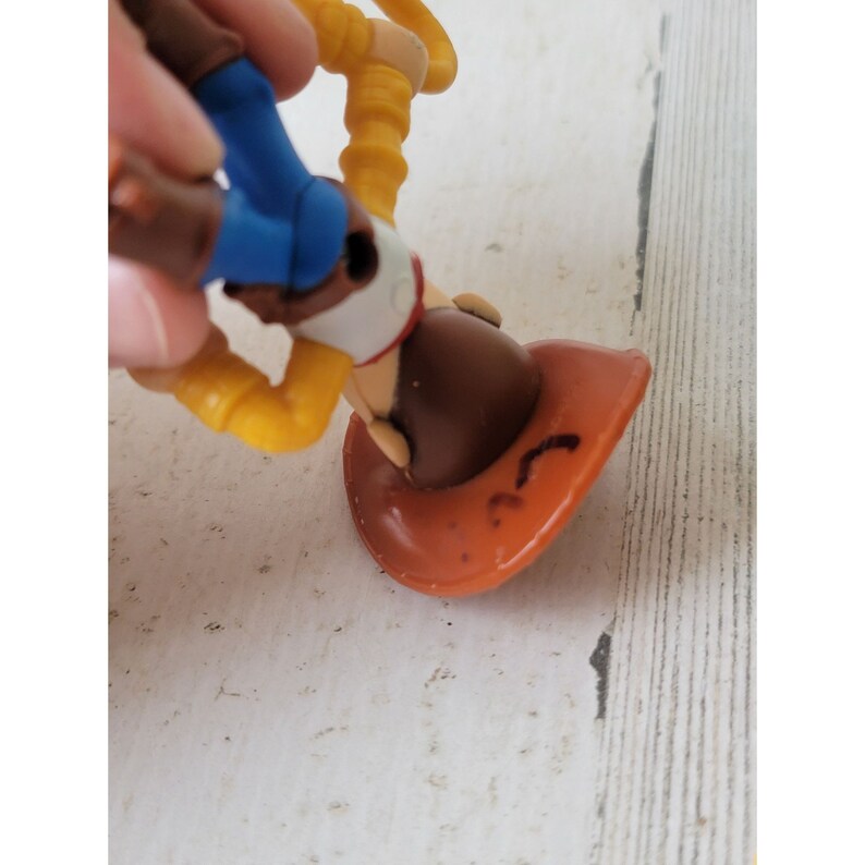 May include: A Woody action figure from the Toy Story franchise. The figure is wearing a brown cowboy hat, a white shirt, a red bandana, a brown vest, blue pants, and yellow sleeves. The figure is being held upside down.