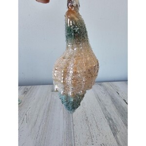 May include: A glass Christmas bauble shaped like a seashell with a white and green iridescent finish. The bauble is covered in small, sparkly beads.