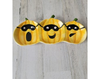 May include: A yellow ceramic serving dish shaped like three pumpkins with black eye masks. The dish is perfect for serving snacks or appetizers.