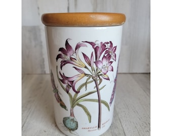May include: White ceramic canister with a wooden lid featuring a botanical illustration of a pink amaryllis flower with green leaves and a butterfly. The text "AMARYLLIS Mexicana" is printed on the bottom of the canister.