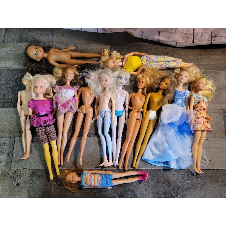 May include: A collection of 12 dolls, including Barbie, Disney Princesses, and other popular characters. The dolls are dressed in a variety of colorful outfits and have different hairstyles.