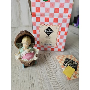 May include: A small fairy figurine sitting inside a walnut shell, with a pink blanket and wings. The figurine is part of the "Little Kitchen Fairies" collection, with the number 1200/21 printed on the box.