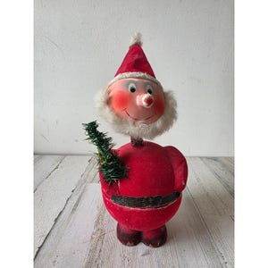 May include: A vintage bobblehead Santa Claus figurine featuring a red velvet-textured body, a white faux fur beard, and a pointed red hat. The figure has a round face with rosy cheeks, a small nose, and holds a miniature green tinsel tree.