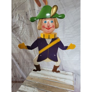 May include: A wooden puppet with a green hat, yellow sash, and purple jacket. The puppet has a painted face with blue eyes and a smile. The puppet is hanging from a string.