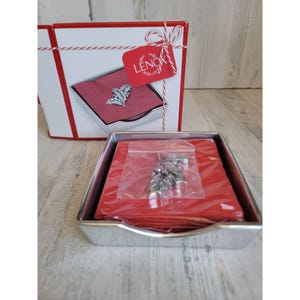 May include: A silver-colored metal napkin holder with a red napkin and a silver leaf-shaped ornament. The ornament is in a plastic bag. A white and red box with a tag that says "LENOX" is in the background.
