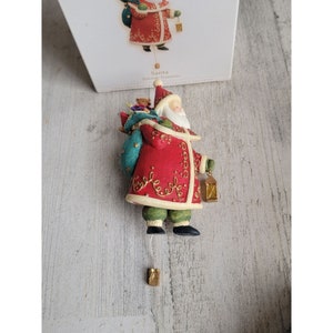 Op de afbeelding: Een rode en gouden kerstman-versiering met een zak vol speelgoed en een lantaarn. De versiering hangt aan een touwtje met een gouden bel aan de onderkant. De tekst "Santa" en "Valued Treasures" is zichtbaar op de doos.