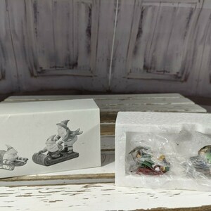 Peut inclure: Deux ensembles de figurines de Noël miniatures dans une boîte blanche. Les figurines sont des lutins qui font du traîneau. Un ensemble est dans un emballage en plastique transparent et l'autre sur un fond blanc.