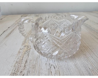 May include: Clear glass creamer with a handle and a spout. The creamer has a cut glass design with geometric patterns. The handle is curved and has a textured design. The creamer is sitting on a light-colored wooden surface.