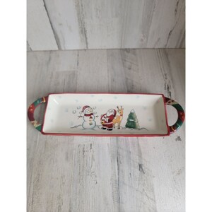 Könnte beinhalten: Eine weiße Keramik-Servierplatte mit rotem und grünem Rand. Die Platte zeigt einen Schneemann, den Weihnachtsmann und ein Rentier in einer Winterszene. Die Platte hat zwei Griffe an den Seiten.