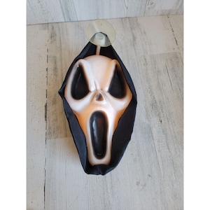 Puede incluir: Una máscara de Halloween de plástico blanco de un fantasma con una túnica negra con capucha. La máscara tiene la boca abierta y una máscara negra para los ojos. La máscara está unida a una ventosa.