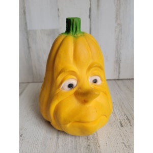 May include: A yellow ceramic pumpkin with a green stem and a cartoon face. The pumpkin has a wide smile, large eyes, and a prominent nose.