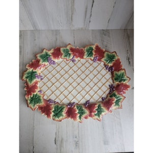 Puede incluir: Un plato de servir ovalado de cerámica con un diseño de celosía elevado en el centro. El borde está decorado con una vid de hojas verdes, hojas rojas y uvas moradas.