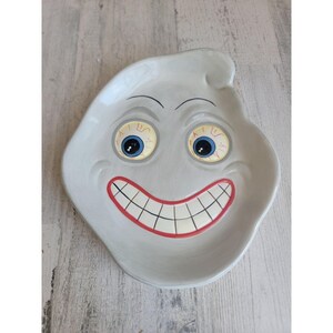 May include: A ceramic ghost-shaped dish with a white glaze and a painted face. The face has red lips, white teeth, blue eyes with black pupils, and red eyebrows.