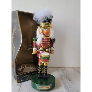 May include: A wooden nutcracker soldier figurine playing a drum. The nutcracker is wearing a red and gold uniform with a white fur trim. The nutcracker is standing on a green base with a "Limited Edition" label. The nutcracker is in a box with the words "Musical Animated Nutcracker Limited Edition".