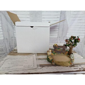 May include: A decorative figurine featuring a miniature farm scene with a fence, tree, and flowers. The figurine has a small, open area, possibly for a photo. A white box and a certificate of authenticity are also present.