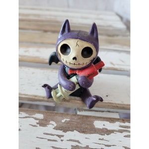 May include: A small purple and white skeletal bat figurine holding a red and white syringe. The figurine has stitched seams and a red bow tie.
