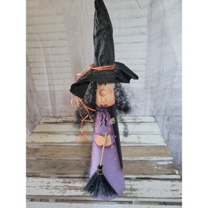 Puede incluir: Una muñeca de bruja morada y negra con un sombrero negro puntiagudo, una cinta naranja y una escoba negra. La muñeca está de pie sobre una superficie de madera blanca.
