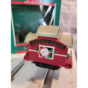 May include: A red and white Christmas caboose ornament with a Santa Claus figure peeking out of the window. The caboose has a gold roof and the words "Christmas Caboose" printed on the side.
