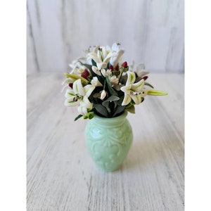 May include: A bouquet of white lilies with green leaves and red accents arranged in a light green ceramic vase.