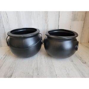 May include: Two black cauldron-shaped pots with rounded bodies and wide rims. One pot has a metal handle. The pots are made of a dark, matte material and are set against a light-colored wooden background. These are ideal for Halloween or themed decor.