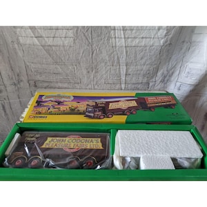 Puede incluir: Un modelo a escala de camión John Codonas Pleasure Fairs Ltd. de Corgi Classics. El camión es marrón con letras blancas y tira de un remolque con la misma marca. La caja es verde y amarilla con una imagen de una feria en la parte delantera.