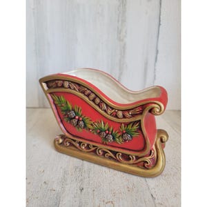 May include: A red ceramic sleigh with a cream-coloured interior, featuring gold-painted scrollwork along the base and rim, and a relief design of green pine needles, brown pinecones, and small red berries along the side of the exterior.
