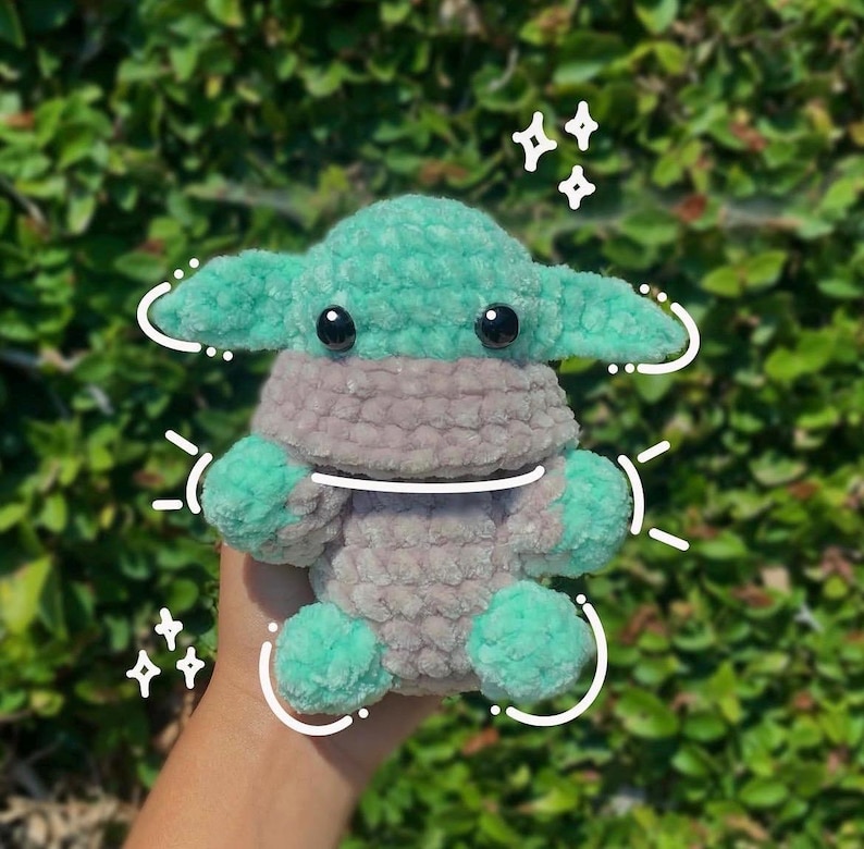 May include: A plush crochet doll featuring a light green head with large black eyes and oversized ears, wearing a tan-coloured garment. The figure is decorated with white hand-drawn digital sparkles and curved lines surrounding the body.