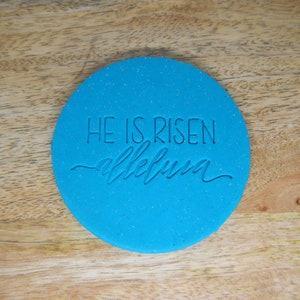 May include: A circular, vibrant blue item with the words "HE IS RISEN" and "alleluia" inscribed. The surface has a speckled appearance, and it's placed on a wooden surface.