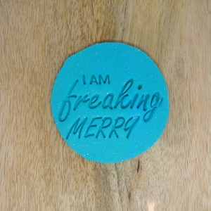 May include: A turquoise blue, oval-shaped cookie cutter with glitter embedded in the surface. The cookie cutter has the text "I am freaking merry" in white.