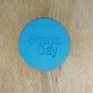 May include: A circular, turquoise coaster featuring the embossed words "Straya Day". The coaster has a speckled appearance and is placed on a light brown wooden surface. The text is in a sans-serif font.