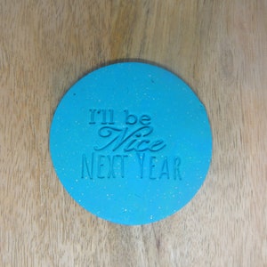 May include: A round blue cookie cutter with glitter embedded in the surface. The cookie cutter has the text "I'll be Nice Next Year" in white.