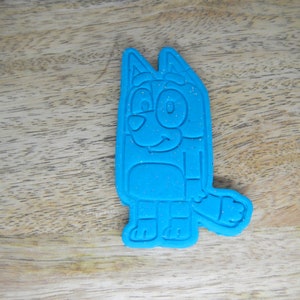 May include: Blue cookie cutter shaped like a cartoon dog with a white eye and a smile. The cutter has a sparkly finish.