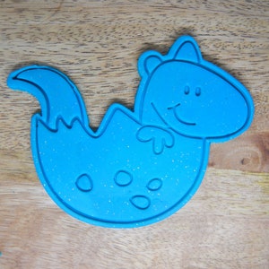 May include: Blue plastic cookie cutter shaped like a dinosaur hatching from an egg. The dinosaur has a smiling face and a small tail.