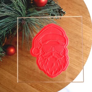 May include: A red plastic Santa Claus head with sunglasses and a beard, on a clear acrylic block.