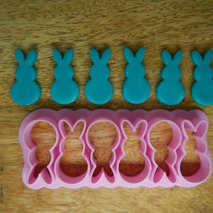 May include: A pink plastic bunny cookie cutter with six individual bunny shapes. Six teal blue bunny-shaped pieces of modelling clay are arranged above the cutter.