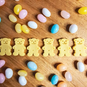 May include: A wooden surface with six yellow gummy bear shaped sweets and a variety of pastel coloured jelly beans.